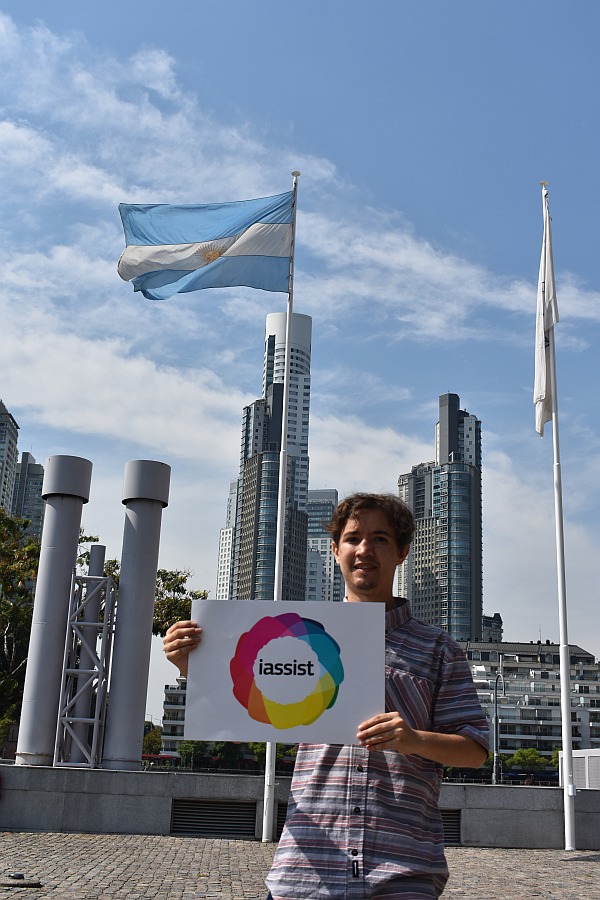 A man standing holding a sign, with Argentinian flag and high rise buildings in the back.