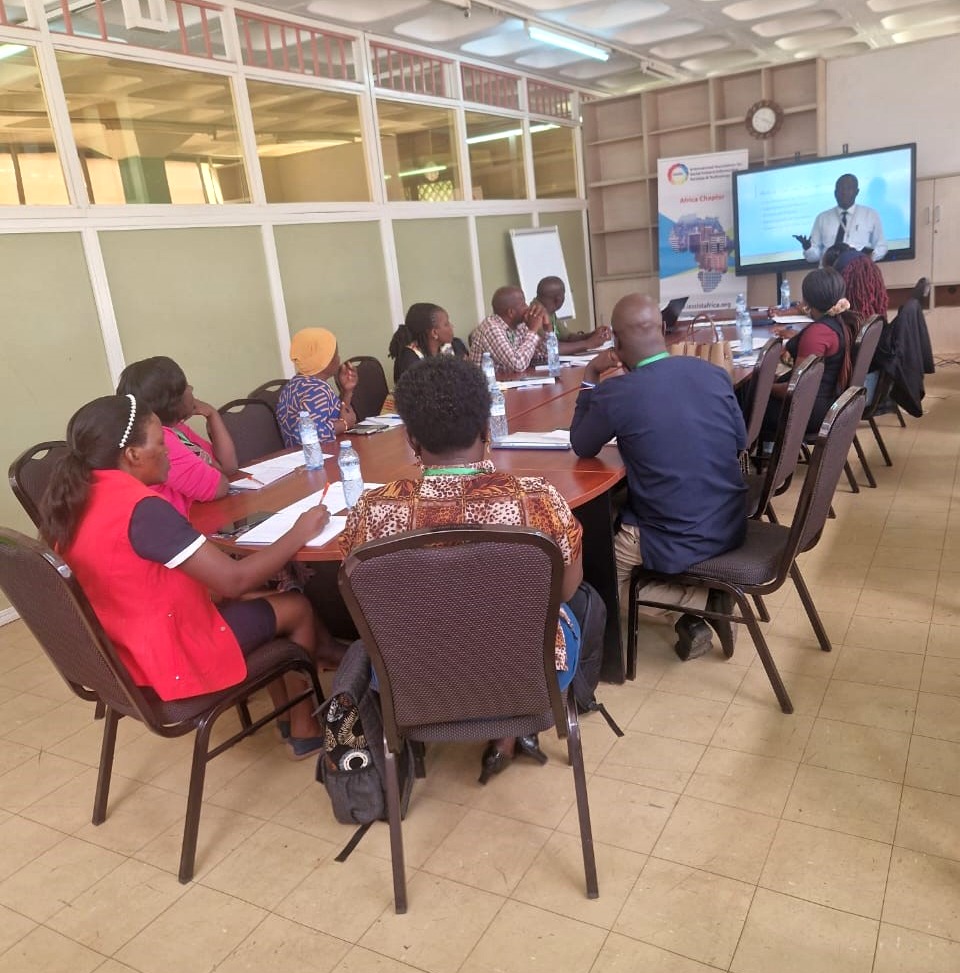 A large group of people sitting around a table, listening to a person standing in front of the large display screen.