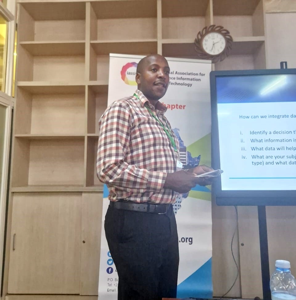A person wearing black trousers and a checkered shirt addressing the audience. There is rollup and large display screen behind the person.