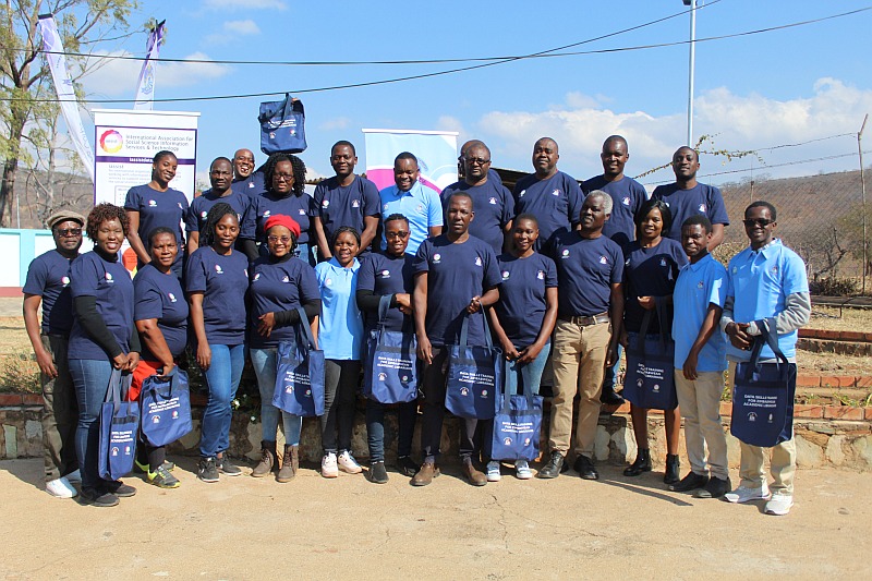 A group of black men and women wearing navy blue or light blue T-shirts and carrying conference bags gathered for a group photo outside in sunny weather.
