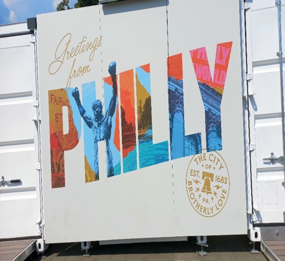 A sign that says Greetings from PHILLY - the city of brotherly love. Philly written in colourful fonts and showing the Rocky statue.